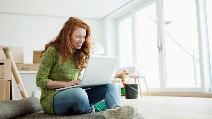 Junge Frau in neuer Wohnung sitzt im Schneidersitz auf dem Boden mit ihrem Laptop, umgeben von Umzugkartons. | © Westend61/Westend61via Getty Images
