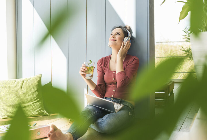 © Westend61/Westend61 via Getty Images Lächelnde Frau mit Kopfhörern und Laptop, die zu Hause am Fenster sitzt | © Westend61/Westend61 via Getty Images