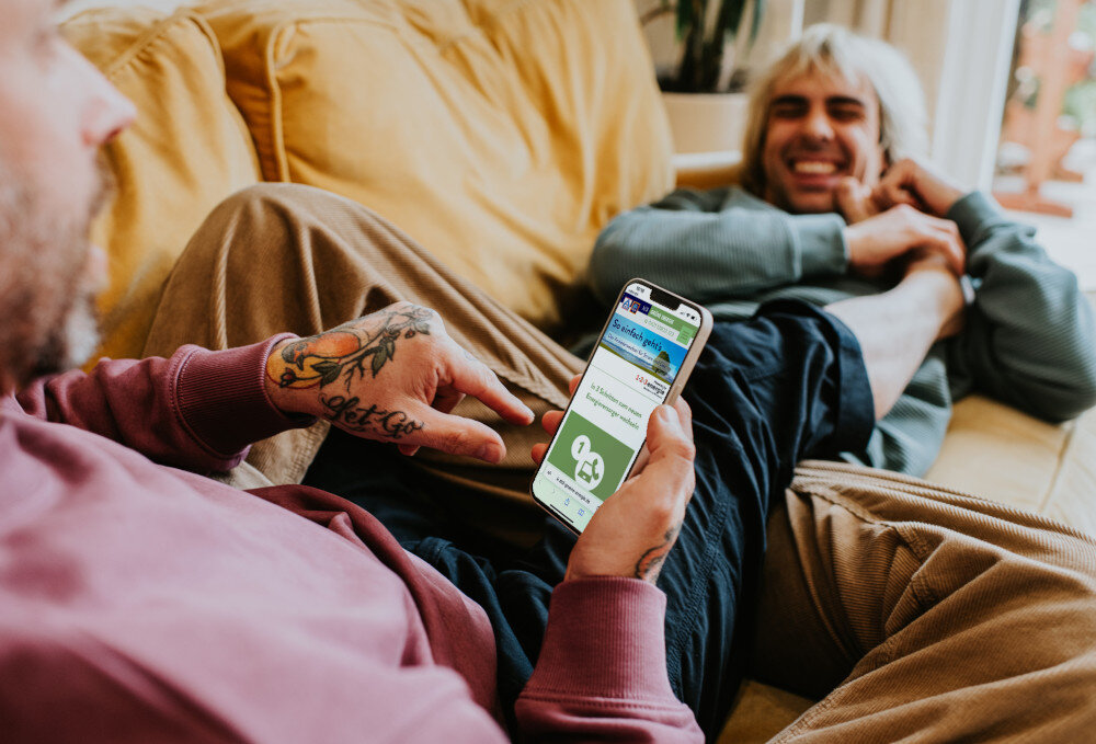 Ein Paar, das auf einem Sofa liegt, steuert seinen Thermostat mit einer Handy-App | © Catherine Falls Commercial/Moment via Getty Images