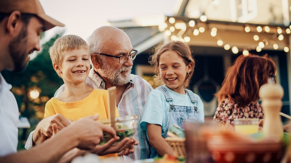 Glücklicher Großvater, der mit seinen Enkelkindern spricht und Spaß hat, sie bei einem Abendessen im Freien mit Essen und Getränken auf dem Schoß hält. Erwachsene auf einer Gartenparty zusammen mit Kindern. Im Hintergrund leuchtet eine Lichterkette. | © © iStock.com | gorodenkoff