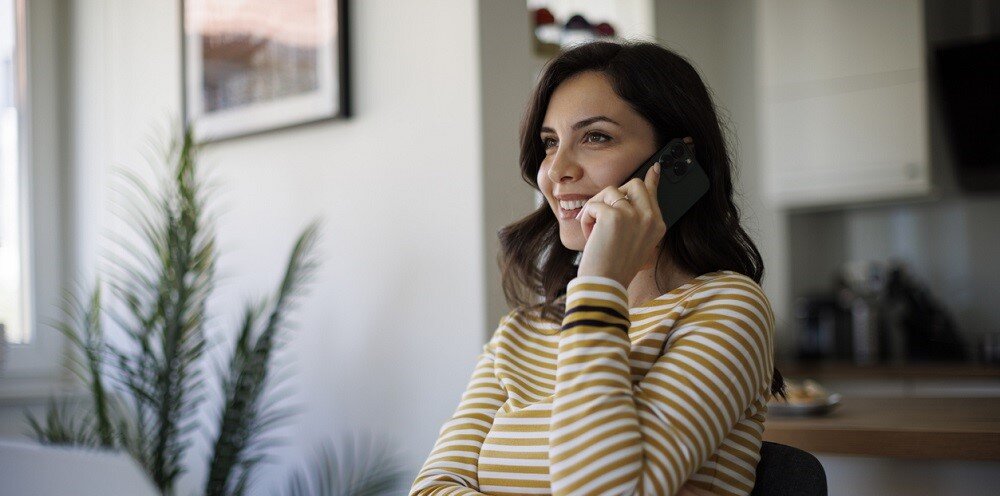 Eine junge Frau sitzt Zuhause an ihrem Laptop und telefoniert mit dem Kundenservice am Handy | © © iStock.com | damircudic