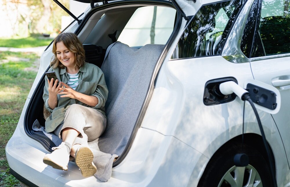 Eine Frau sitzt im geöffneten Kofferraum ihres Elektroautos und lächelt freudig auf ihr Smartphone-Display, während das Auto geladen wird. | © © iStock.com | Scharfsinn86